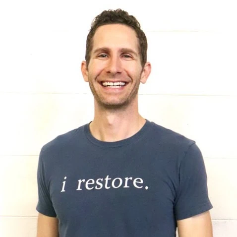 Smiling man wearing "i restore." t-shirt standing in front of a white wall.